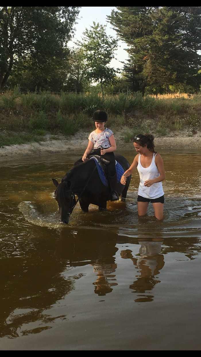 La monitrice accompagne une jeune sur son cheval pour traverser la rivière lors d'une colonie de vacances d'équitation