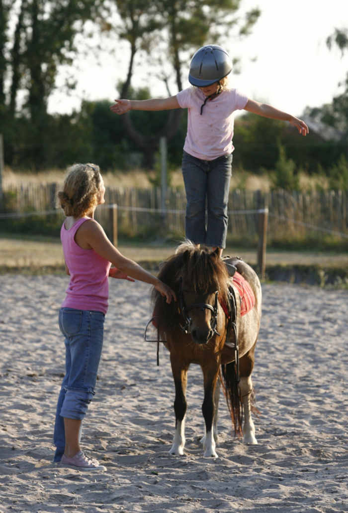 La monitrice aide une fille a se tenir debout sur son poney lors d'une colonie de vacances d'équitation