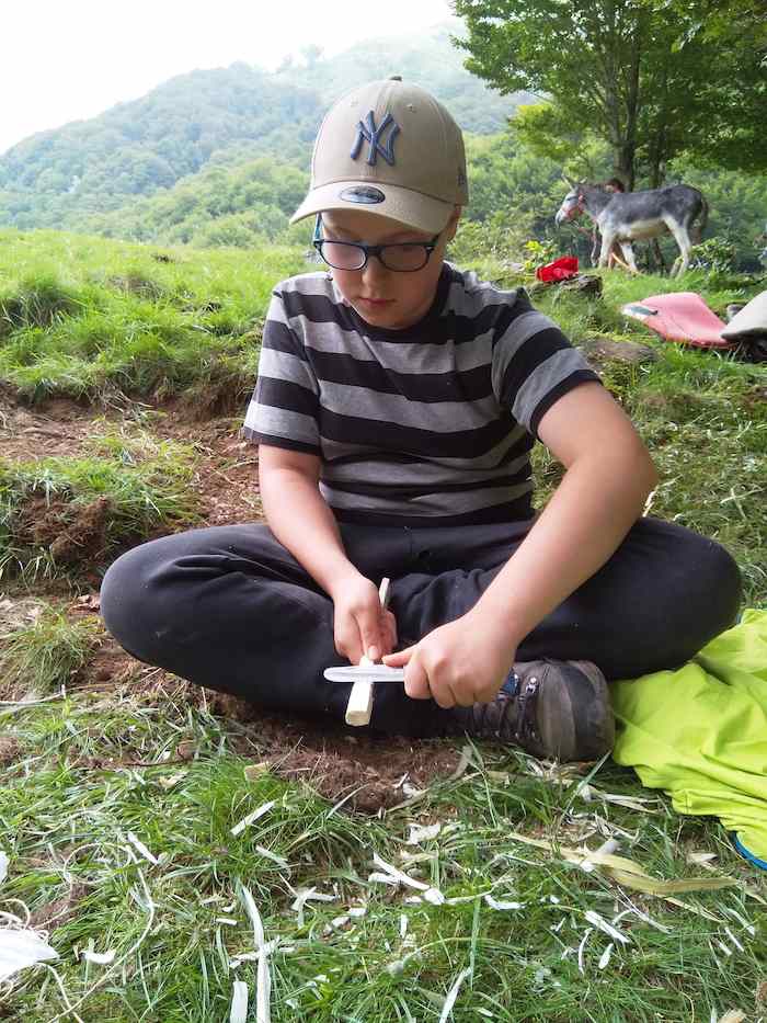 Un enfant sculpte un bout de bois assis dans l'herbe lors d'une colonie de vacance nature dans les Pyrénées