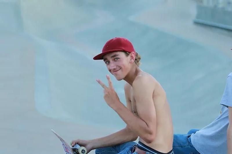 Jeune garçon qui fait un signe peace torse nu avec une casquette rouge avec un skate dans l'autre main assis en stage skateboard