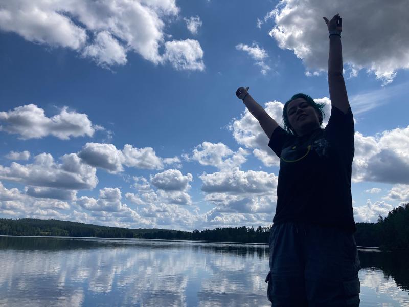 Jeune personne les bras levés pour la photo avec le ciel bleu et les nuages se reflétant sur le lac, pendant le camp itinérant en Suède