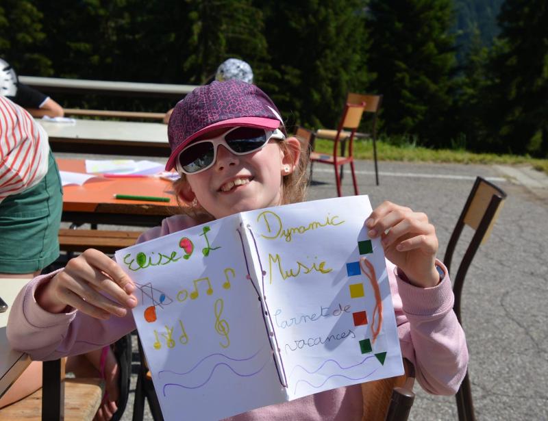 enfants avec une casquette et des lunettes de soleil qui nous montre son carnet de vacances de musique préparé en colo